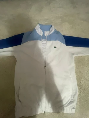 Vit och blå kofta/sommarjacka från Lacoste - Snygg sommarjacka från Lacoste i vitt med ljusblå och mörkblå detaljer över axlar och ärmar. Jackan har dragkedja framtill, ståkrage och två fickor. Klassisk Lacoste-logga på bröstet. Perfekt för en sportig och stilren look.