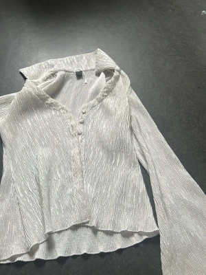 Vit plisserad blus med krage - Säljer en vit plisserad blus med lång ärm och klassisk krage. Blusen har knappar framtill och ett lätt glansigt tyg som ger en trendig look. Perfekt för dig som gillar stilrena och moderna plagg med en twist.