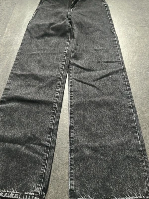 Svarta raka jeans  - Säljer ett par svarta jeans från H&M med rak passform och hög midja. Jeansen har klassisk femficksdesign, dragkedjegylf och är tillverkade i denim med lätt tvättad look. Perfekta för en avslappnad och trendig stil.