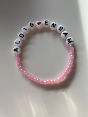 Pärlarmband med text 'Aldrig Ensam' - Sött armband med rosa pärlor, där de vita pärlorna bildar texten 'Aldrig Ensam' och har ett litet rött hjärta. Perfekt accessoar för att sprida pepp och kärlek. Elastiskt band gör det enkelt att ta på och av.