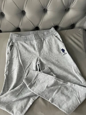 Gråa mjukisbyxor från US polo barn - Snygga gråa mjukisbyxor från US polo med blå logga broderad på vänster ben. Byxorna har resår i midjan, muddar vid bensluten och två sidofickor. Tillverkade i mjukt bomullsmaterial som är perfekt för chill dagar. Storlek 11-12 år eller 146-152 cm