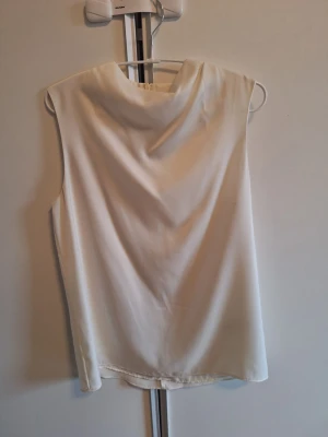 Vit ärmlös linnetopp med draperad hals - Säljer en stilren vit linnetopp med draperad halsringning. Toppen är ärmlös och har en mjuk, något glansig yta som ger ett elegant intryck. Perfekt att matcha med jeans eller kjol för en clean look.