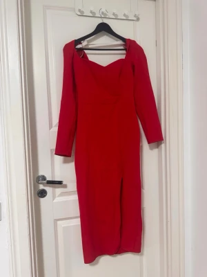 Röd långärmad klänning med slits - Snygg röd långklänning med långa ärmar och en elegant slits framtill. Klänningen har en v-ringad urringning och figurnära passform som framhäver siluetten. Perfekt för dig som vill sticka ut med en klassisk och trendig look.