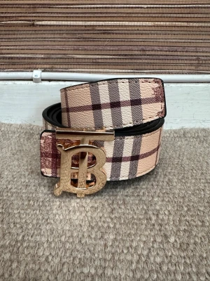 Rutig Burberry bälte med guldspänne - Snyggt bälte från Burberry med klassiskt rutigt mönster i beige, brunt och vitt. Bältet har ett stort guldigt spänne med Burberrys ikoniska monogram. Perfekt accessoar för att lyfta din outfit.
