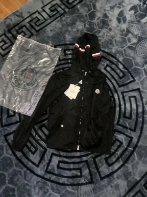 Svart vindjacka från Moncler - Svart vindjacka från Moncler med huva och dragkedja framtill. Jackan har vita och röda ränder på huvan samt Moncler-logga på bröstet. Lätt material, perfekt för blåsiga dagar. Snygg och sportig design med fickor och justerbar nederkant.