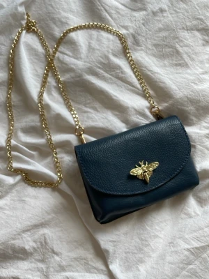 Mörkblå kuvertväska med guldkedja - Snygg mörkblå kuvertväska i läderimitation med guldfärgad kedja och ett coolt guldfärgat bi som detalj på locket. Perfekt storlek för mobilen och det viktigaste. Enkel att bära crossbody eller på axeln. Köpte i italien för 450 kr. 