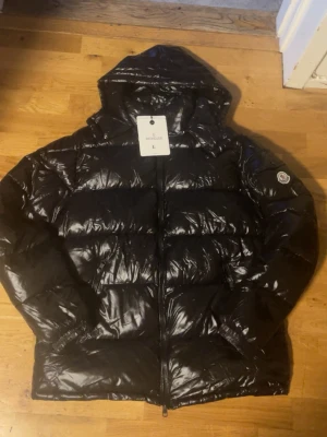 Svart dunjacka från Moncler - Snygg svart dunjacka från Moncler med glansig finish och klassisk logotyp på ärmen. Jackan har dragkedja framtill och två fickor. Perfekt för kalla vinterdagar och riktigt trendig stil.