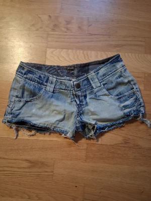 Ljusa jeansshorts med fiskbrodyr - Säljer ett par ljusa, slitna jeansshorts med råa kanter och coola slitningar framtill. På baksidan finns en stor broderad fisk i orange och vitt som ger en unik touch. Shortsen har klassiska fickor och bälteshällor.