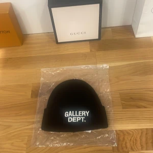 Svart mössa från Gallery Dept. - Helt ny och oanvänd Snygg svart mössa från Gallery Dept. med vit text på framsidan. Perfekt för att hålla stilen i kallare väder. Passar till både vardag och festliga tillfällen. Enkelt att matcha med olika outfits.