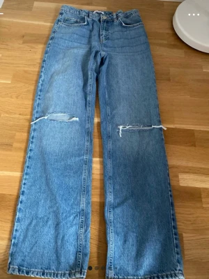 Blå raka jeans med slitna knän - Säljer ett par blå raka jeans i klassisk denim med slitna detaljer över båda knäna. Jeansen har hög midja, fem fickor och stängs med dragkedja och knapp. Perfekta för en avslappnad och trendig look.