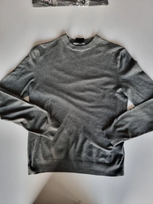 Grå stickad tröja med rund hals - En stilren grå stickad tröja med rund halsringning och långa ärmar. Tröjan har ribbade muddar vid ärmslut och nederkant för en snygg passform. Perfekt basplagg i mjukt material som passar till det mesta.