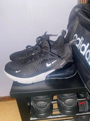 Nike Air Max 270 svarta sneakers - Svarta Nike Air Max 270 sneakers med vit sula och synlig Air-enhet i hälen. Ovandelen är i mesh för bra andningsförmåga och har vita detaljer som Swoosh-loggan och texten 'AIR270'. Skorna har snörning och en sportig, modern look.