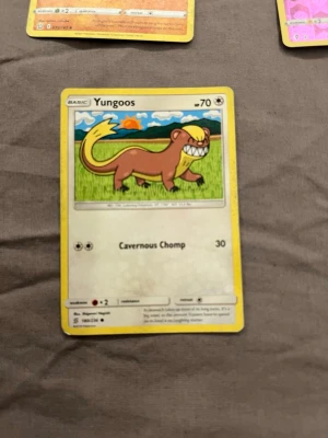 Pokémonkort Yungoos - Pokémonkort med motivet Yungoos. Kortet har gul ram och visar en tecknad figur mot en grön äng med blå himmel och sol. Texten 'Cavernous Chomp' och siffran 30 finns på kortet. Perfekt för samlare eller Pokémonfans.