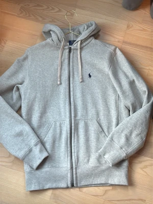 Grå hoodie med dragkedja från Polo Ralph Lauren - Ljusgrå hoodie med dragkedja från Polo Ralph Lauren. Klassisk design med huva och dragsnören, två fickor framtill och broderad logga på bröstet. Mjuk bomullsblandning som är perfekt för en avslappnad stil. Och är såklart äkta och helt ny bara tvättad en gång