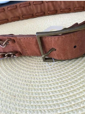 Brunt bälte i mocka med kedjedetaljer - Snyggt brunt bälte i mocka med coola kedjedetaljer längs kanterna och ett spänne i metall. Perfekt accessoar för att lyfta din outfit med en touch av vintage och edge.