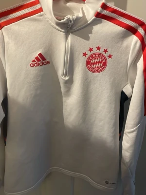 Vit FC Bayern München träningströja - Snygg vit långärmad träningströja från Adidas med FC Bayern München-logga och röda detaljer. Halvlång dragkedja framtill, röda ränder på axlarna och svart/röda inslag på ryggen. Perfekt för fotbollsträning eller sportiga dagar.