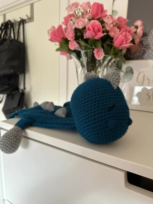 Virkad snuttefilt drake - Virkad drake i form av en snuttefilt. Den är gjord i akrylgarn i färgen petroleumblå med grå detaljer. Den är ungefär 40 cm lång. 