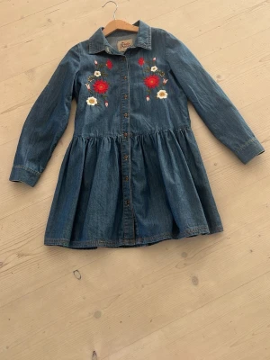 Jeansklänning med broderade blommor - Snygg jeansklänning med knappar framtill och broderade blommor i rött och vitt på bröstet. Klänningen har lång ärm, klassisk krage och utställd kjol.