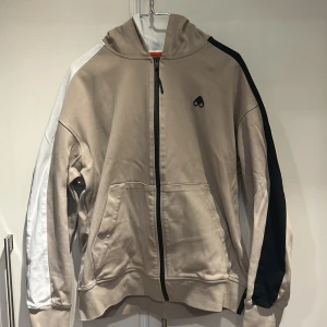 Beige hoodie från Moose Knuckles - Snygg beige hoodie från Moose Knuckles med dragkedja framtill, svarta och vita detaljer på ärmarna samt logga på bröstet. Huva med tryckt text och två fickor framtill. Perfekt för en avslappnad streetstyle.