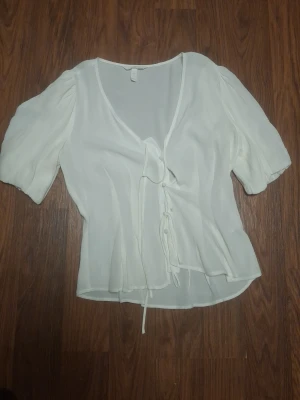 Vit blus med knytband och puffärm - Söt vit blus i tunn, luftig chiffong med v-ringning och knytband framtill. Blusen har korta puffärmar och en lätt utsvängd passform som ger en romantisk känsla. Perfekt att styla med jeans eller kjol för en trendig look.