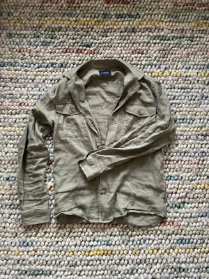 Olivgrön overshirt från Bondelid - Stilren grön overshirt från Bondelid i mjukt linnematerial. Skjortan har två bröstfickor med knappar, klassisk krage och långa ärmar. Perfekt lager-på-lager-plagg för en avslappnad look.