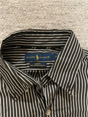 Svartvit randig skjorta från Ralph Lauren - Säljer en slim fit skjorta från Ralph Lauren i svart och vitt med klassiskt randigt mönster. Skjortan är tillverkad i 100% bomull stretch och har knappar framtill samt långärmad design. Perfekt för dig som gillar stilrena och tidlösa plagg.