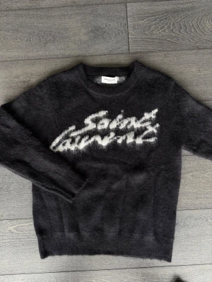 Svart Saint Laurent stickad tröja - Cool svart stickad tröja från Saint Laurent med vit logotyp i grafisk stil på bröstet. Tröjan har rund hals och långa ärmar, och är gjord i ett fluffigt material som ger en lyxig känsla. Perfekt för dig som vill ha något trendigt och exklusivt.