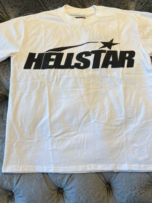 Vit t-shirt från Hellstar - Vit t-shirt från Hellstar med stor svart logga och stjärndetalj på bröstet. Klassisk passform och rund halsringning, perfekt för en avslappnad streetwear-look.