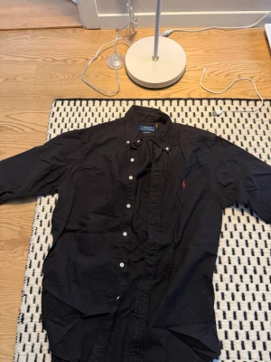 Svart skjorta från Polo Ralph Lauren - Klassisk svart skjorta från Polo Ralph Lauren med knappkrage och vita knappar. Skjortan har lång ärm och den ikoniska röda logotypen broderad på bröstet. Perfekt till jeans eller chinos för en clean look.