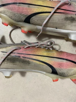Puma ultra färgglada fotbollsskor - Säljer ett par Puma ultra fotbollsskor med en snygg mix av rosa, gul och svart på ovandelen. Skorna har en låg profil, snörning och slitstarka dobbar i orange och vitt. Tillverkade i syntetmaterial för bästa känsla på planen.