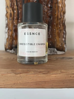 ESSNCE Irresistible Enigma parfym - En stilren parfymflaska från ESSNCE med doften Irresistible Enigma. Flaskan är cylindrisk i genomskinligt glas med svart matt kork och vit etikett med svart text. Innehållet är gyllengult och flaskan har en modern, minimalistisk design.