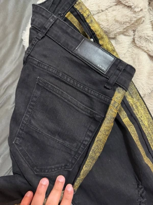 Svarta jeans med gulddetalj från Amiri - Säljer ett par svarta jeans från Amiri med unika guldfärgade ränder längs sidorna. Limited jeans. Köptes i marbella finns nån enstaka som säljer för 6000+ på eBay etc. Authentic model number finns på tag.