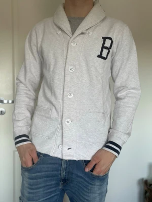 Ljusgrå cardigan med B-detalj - Snygg ljusgrå cardiganjacka med stora vita knappar. Från märket bondeid. En liten defekt på kragen(bild 4) samt lite nopprig. 