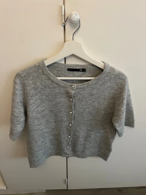 Grå kortärmad kofta från 157 - Snygg grå stickad kofta från 157 med korta ärmar och rund halsringning. Koftan har pärlknappar framtill och ribbade kanter. Perfekt att slänga över en topp för en chill look.