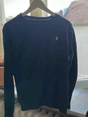 Mörkblå stickad tröja från U.S. Polo Assn. - Mörkblå stickad tröja från U.S. Polo Assn. med klassisk rund halsringning och broderad logga i vitt på bröstet. Tröjan har långa ärmar och ribbade muddar vid ärmslut och nederkant. Perfekt för en stilren och avslappnad look.