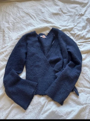 Mörkblå omlottkofta med vida ärmar - Säljer en mörkblå omlottkofta med långa, vida ärmar och mjukt, fluffigt material. Koftan har knytband i sidan för att justera passformen och en avslappnad, trendig look. Perfekt för lager på lager och enkel att styla till olika outfits.