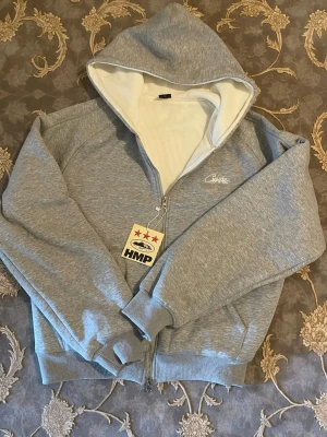 Corteiz hoodie - Snygg grå zip hoodie från HMP med vit insida i huvan och broderad logga på bröstet. Dragkedja framtill och en klassisk passform. Perfekt för en chill och avslappnad stil. Hoodien består 100% av bomull samt att priset går även att diskuteras.