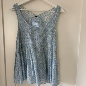 Blåvit ärmlös blommig topp Bondelid. - Supersöt ärmlös linnetopp från Bondelid med blått och vitt blommönster. Toppen har knytning i ryggen och en luftig, flowy passform. Perfekt för varma dagar och enkel att styla med jeans eller kjol. Längd 63 cm.