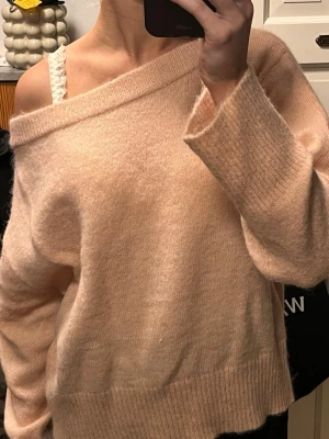 Ljusrosa off shoulder stickad tröja - Mysig ljusrosa stickad tröja med off shoulder-design och bred ribbad kant vid halsen. Tröjan har en avslappnad passform och långa ärmar. Perfekt för dig som gillar en trendig och bekväm stil.