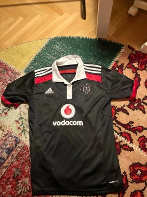 Orlando Pirates matchtröja Adidas - Snygg svart matchtröja från Orlando Pirates med vita och röda detaljer. Tröjan har klubbmärke på bröstet, vit krage och Adidas-logga. Tillverkad i lätt och ventilerande material, perfekt för fotboll eller träning. Sponsrad av Vodacom.