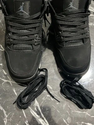 Svarta Air Jordan 4 sneakers - Säljer ett par svarta Air Jordan 4 sneakers med klassisk Jumpman-logga på tungan. Skorna har meshpaneler, robust sula med synlig luftdämpning och extra skosnören. Materialet är en mix av läder och syntet. Perfekt för dig som vill ha en ikonisk och stilren sneaker.