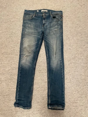 Blå jeans från Replay med slitningar - Säljer ett par blå jeans från Replay med snygga slitningar och tvättade detaljer på låren och knäna. Klassisk femficksmodell med knappgylf och raka ben. Perfekta för en avslappnad och trendig stil.