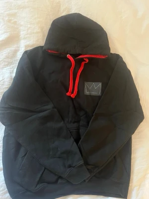 Svart Hov1 Hoodie  - Svart hoodie med huva.På bröstet finns en vit krona tryckt och på ryggen en stor patch med texten 'VINDAR TILL MARS' i färgglada bokstäver. Hoodien har känguruficka och är gjord i mjukt bomullsmaterial. 