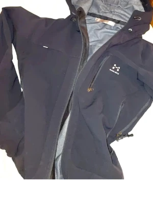 Svart vindjacka från Haglöfs - Snygg svart vindjacka från Haglöfs med huva och dragkedja. Jackan har flera praktiska fickor med dragkedja och justerbar nederkant med dragsko. Perfekt för blåsiga dagar och outdoor-aktiviteter. Materialet är slitstarkt och vattenavvisande. Kan sänka prisen vi kan diskuteras 