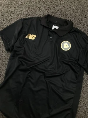 Svart pikétröja från New Balance - Svart pikétröja från New Balance med guldiga loggor på bröstet. Tröjan har klassisk krage, knappar framtill och är kortärmad. Perfekt för en sportig och stilren look.