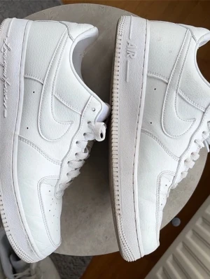 Nike Air Force 1 Drake Nocta ”Certified lover boy” Storlek 44 - Klassiska Nike Air Force 1 sneakers i helvitt läder med vit sula och matchande vita skosnören. Ikonisk swoosh-logga på sidorna och präglad AIR-text på sulan. Stilren och tidlös modell som passar till många outfits. En extra uppsättning vita skosnören och blåa bokstavspärlor medföljer. Skorna har slitage på insidan av hälkappan och smuts på sulorna. Storlek 44 pris kan diskuteras