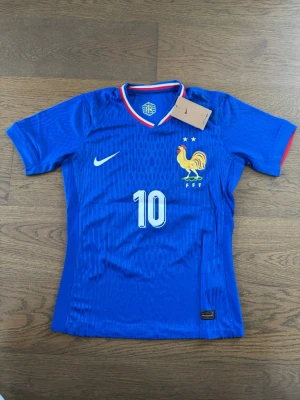 Frankrike Mbappé matchtröja blå - Blå fotbollströja från Nike med Frankrikes landslagslogga och nummer 10 på framsidan. Mbappé och nummer 10 tryckt på ryggen. Tröjan har korta ärmar, röd och vit detalj vid kragen och är tillverkad i lätt, ventilerande material.