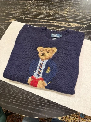 Mörkblå stickad tröja Polo Bear - En mörkblå tröja med en stor, detaljerad teddybjörn på framsidan. Björnen är klädd i en blå kavaj med guldiga knappar och ett emblem, en vit skjorta med en randig slips, samt röda byxor och bruna skor. Tröjan har en rund halsringning och ribbade muddar vid ärmslut och nederkant. Storleken är small men passar till Medium pris kan diskuteras 