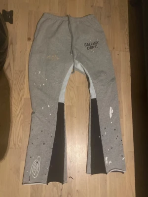 Grå mjukisbyxor Gallery Dept. - Unika grå mjukisbyxor från Gallery Dept. med svart och vitt färgstänk, svarta paneler längs benen och tryckt logga på ena benet. Byxorna har elastisk midja och raka ben med slits nertill. Perfekt för dig som gillar streetwear och en kreativ look.
