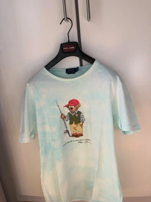 Polo Ralph Lauren: Polo Bear TieDye Limited Edition T-Shirt - Denna fantastiska TieDye T-Shirten från Polo Ralph Lauren är en Limited Edition Polo Bear T-Shirt med en fiskare som version av Polo Bear. Säljer denna som är nästan som helt ny då den är för stor för mig (Stor XL) 🎉 Tröjan är som helt ny och köpt på Boozt för 1995 kronor. 🎉För mer info skriv dm!